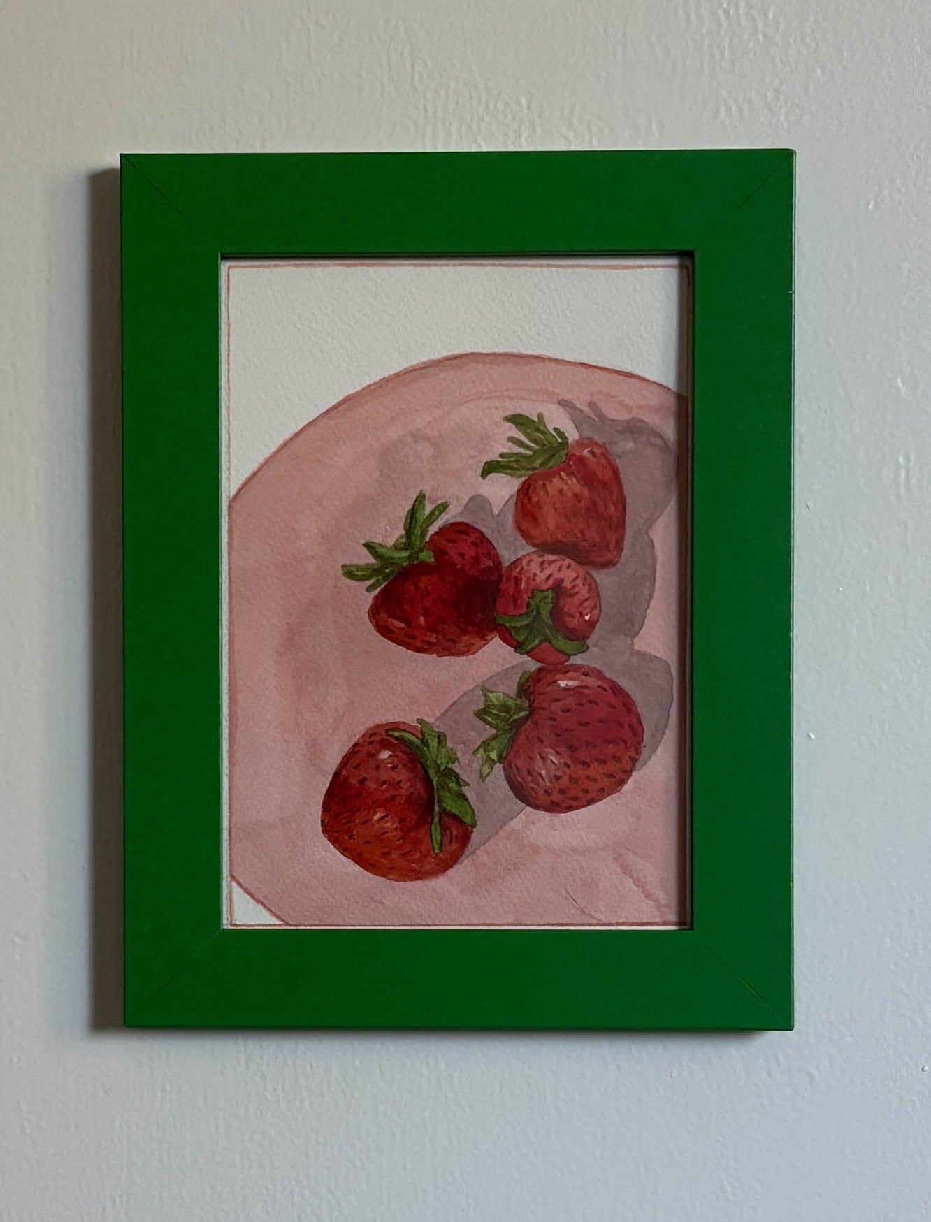 A framed still life painting of strawberries on a pink plate, hung on a textured off-white wall. The artwork features a vibrant green frame.