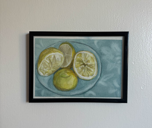 A framed still life painting of lemons on a plate, hung on a textured off-white wall. The painting features yellow lemons on a light blue-gray plate against a muted blue-gray background, enclosed in a black frame with a white mat.