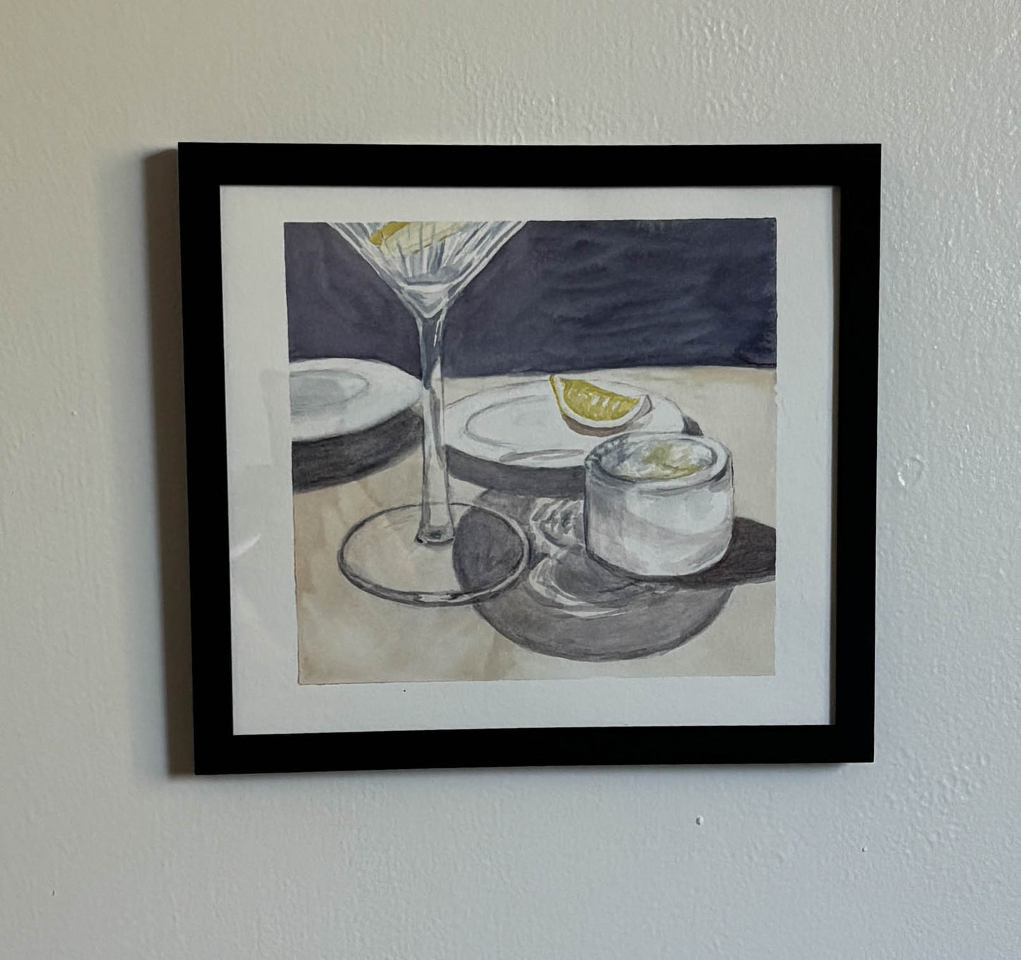 A framed watercolor painting of a martini glass, a small ice bucket, and a lemon slice, displayed on a textured white wall.
