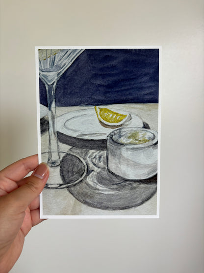 A hand holds a printed art card featuring a still life illustration of a martini glass, a plate with a lemon wedge, and a small glass with a drink, against a neutral light gray background.