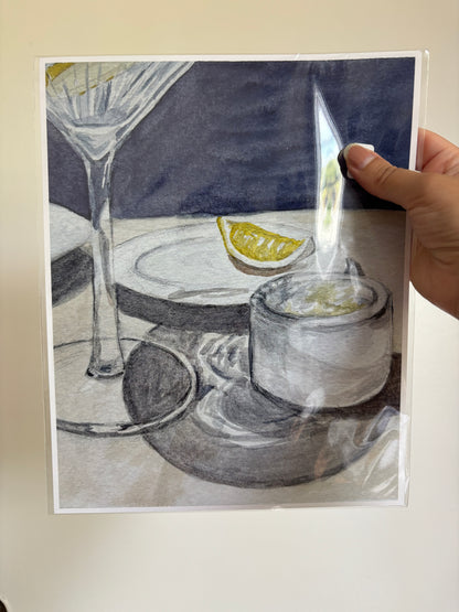 A hand holds a square art print or card featuring a still life drawing of a martini glass, a small cup, and a lemon wedge on a dark blue background. The print is photographed against a plain light wall, with a glare reflection visible on the plastic sleeve.