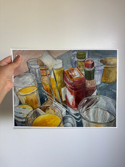 A hand holds a square watercolor painting of various glass containers, bottles, and jars on a table, shown against a plain off-white wall.