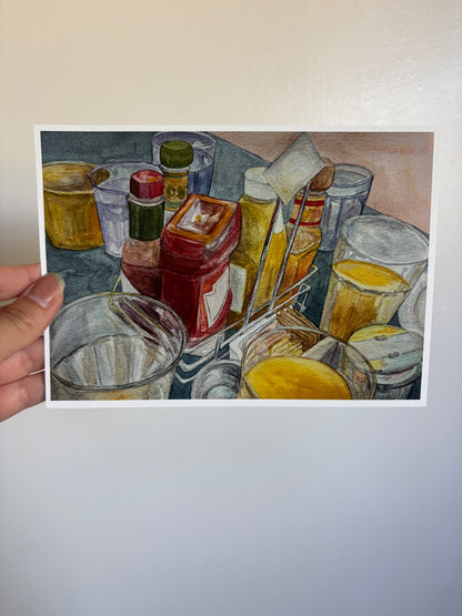 A hand holds a printed artwork showing a still life of glassware, bottles, and yellow liquid, photographed against a plain light wall.