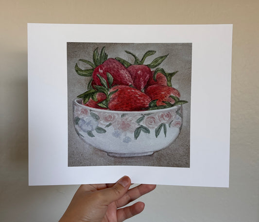 A hand holds a white art print featuring a still life illustration of red strawberries in a floral white bowl, set against a neutral beige wall.
