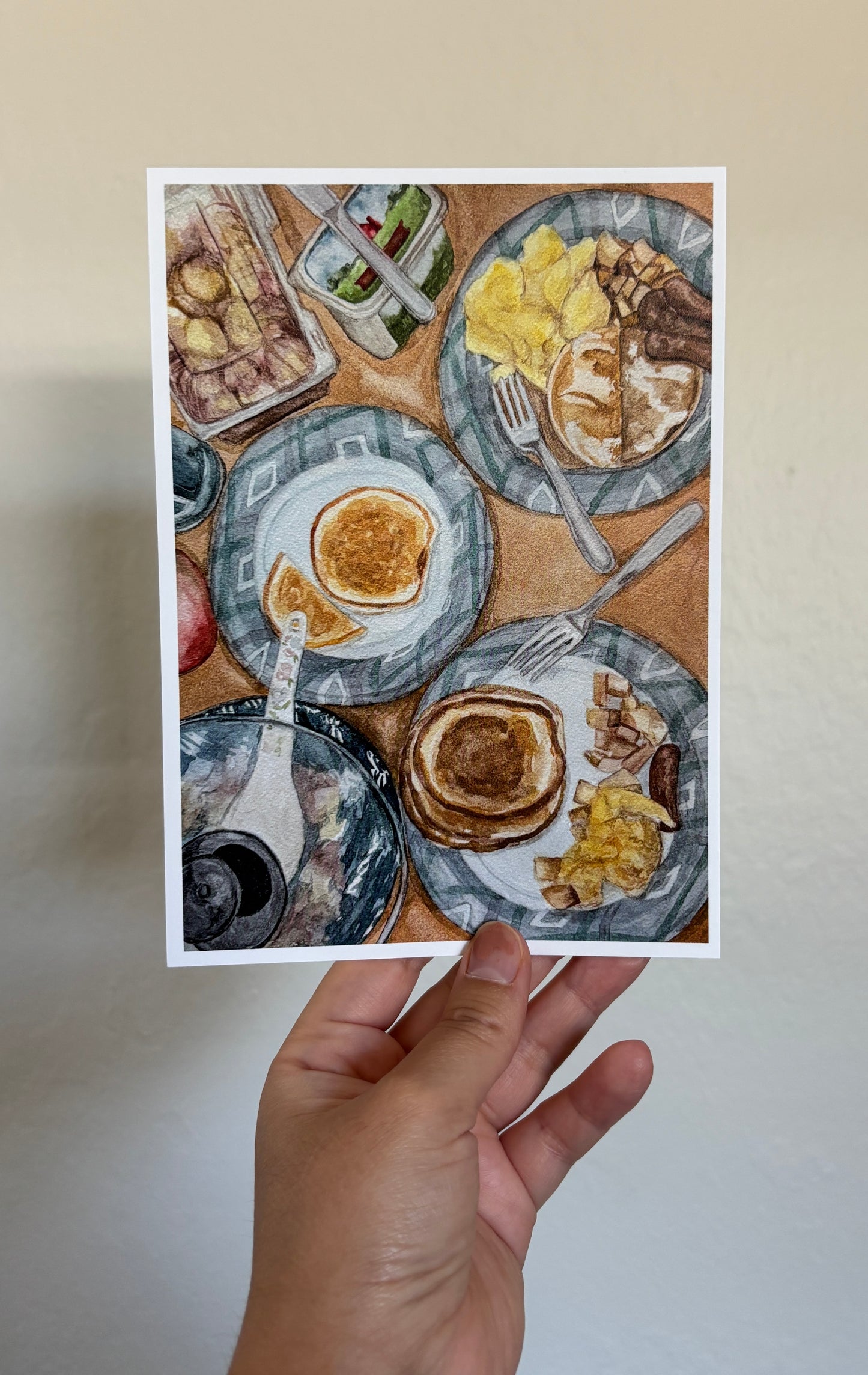 A hand holds a square art print featuring a watercolor-style breakfast table scene with pancakes, eggs, bacon, and plates, against a plain beige wall.