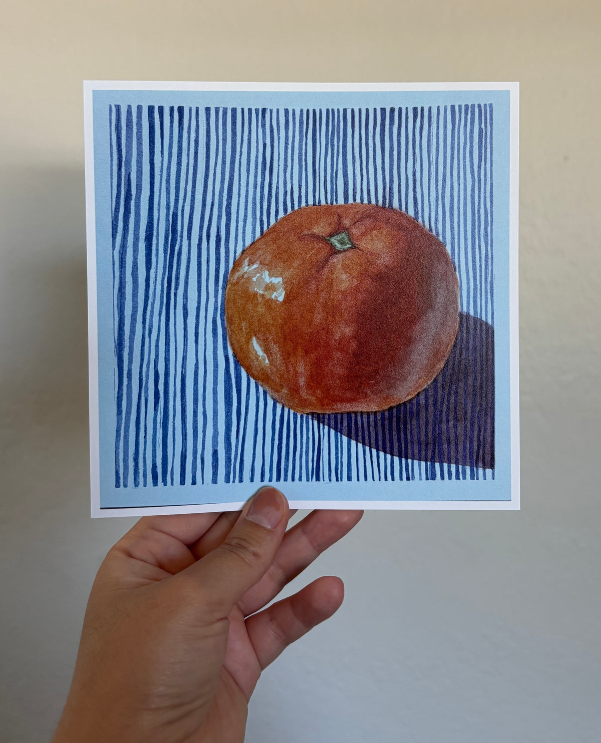 A hand holds a square art print featuring a painted orange on a blue striped background, against a plain beige wall.