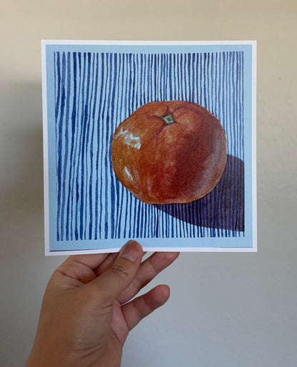 A hand holds a square art print featuring a painted orange on a blue striped background, against a plain beige wall.