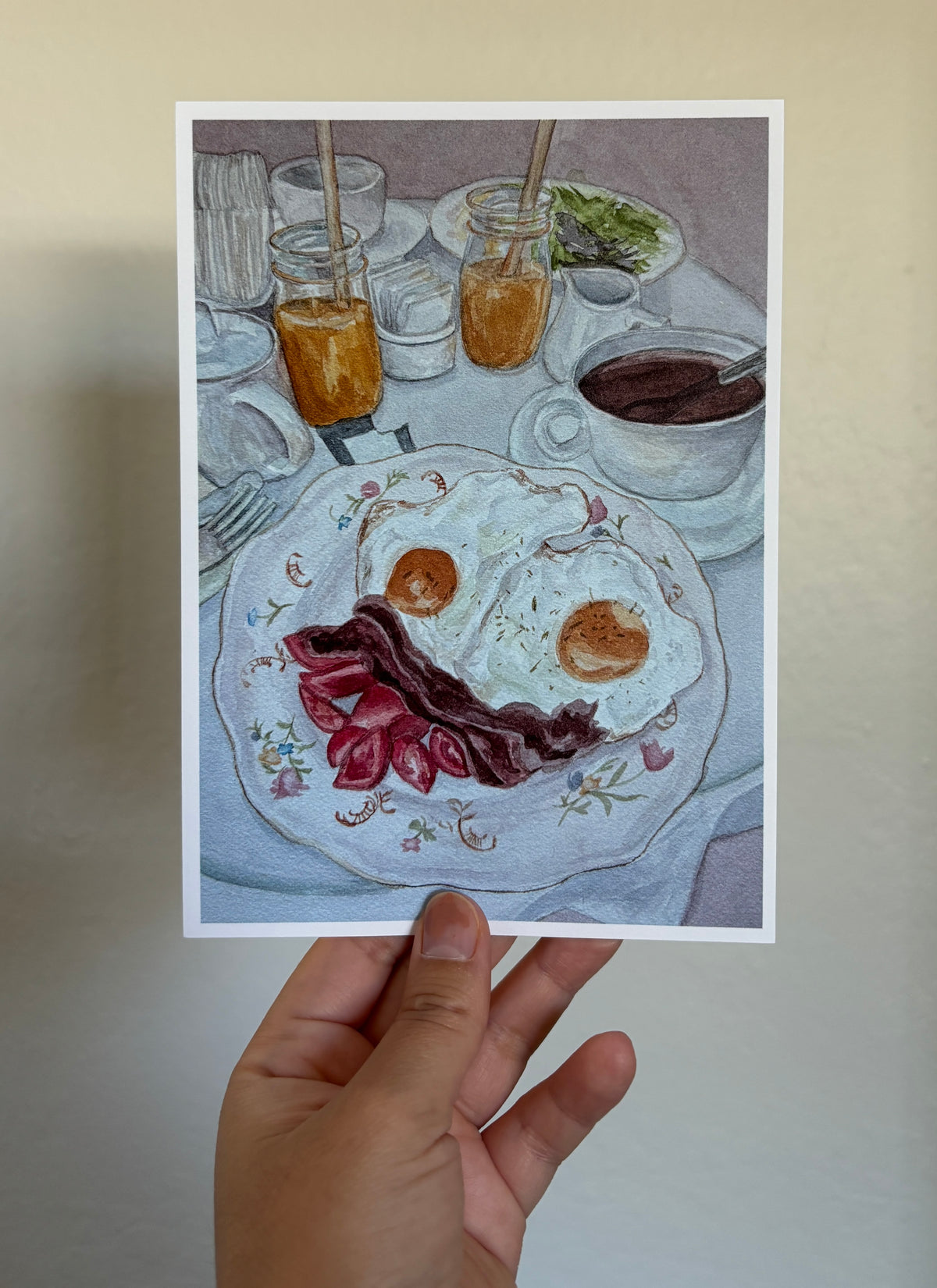A hand holds a square art print depicting a breakfast table scene with a plate of eggs, bacon, and sausage, mason jars with orange drinks, and a cup of coffee, against a plain beige wall.