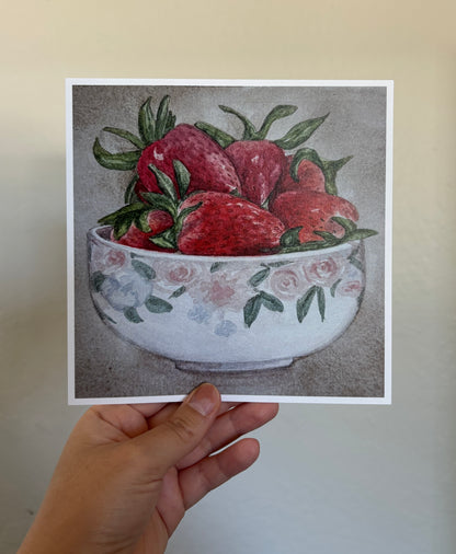 A hand holds a square art print featuring a still life illustration of red strawberries in a white floral bowl, against a neutral beige wall.