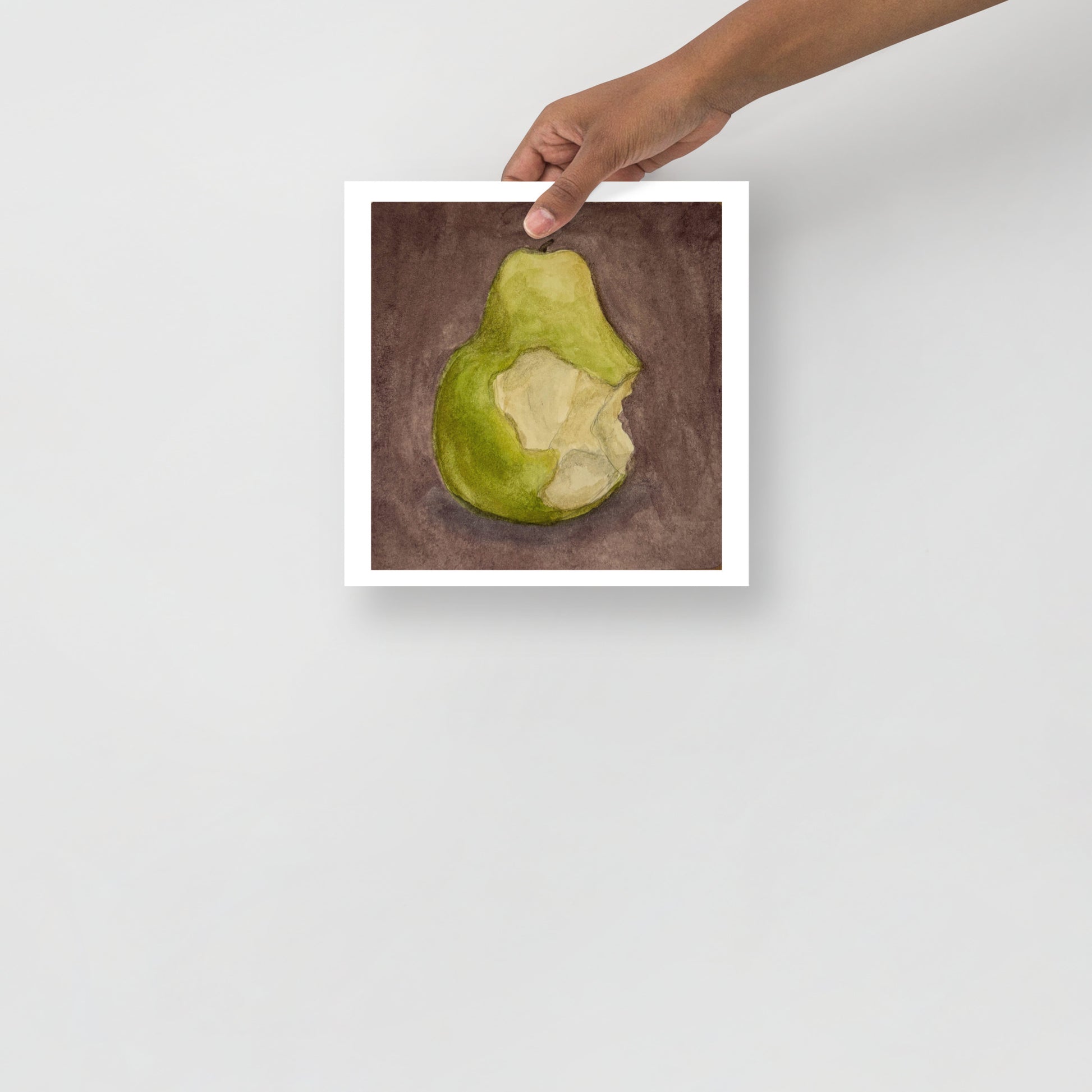 Product mockup image includes a hand holding a print of my painting of a green pear with a bite taken out of it against a plum colored background. 