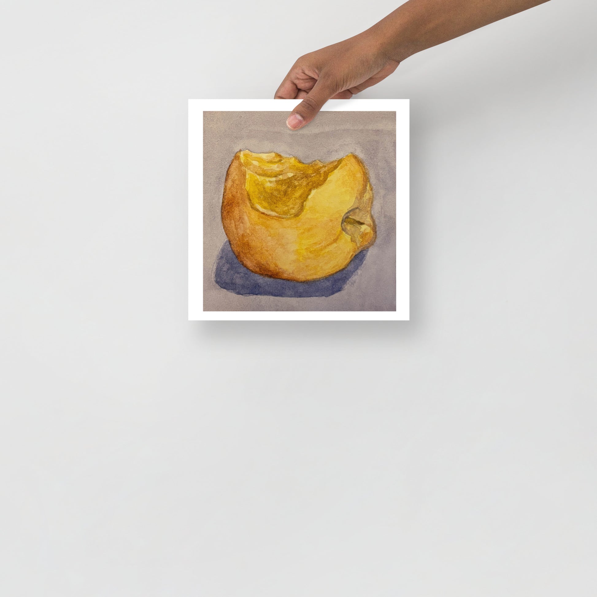 Product mockup of a watercolor painting that depicts a golden apple with a bite taken out of it against a greyish background. 