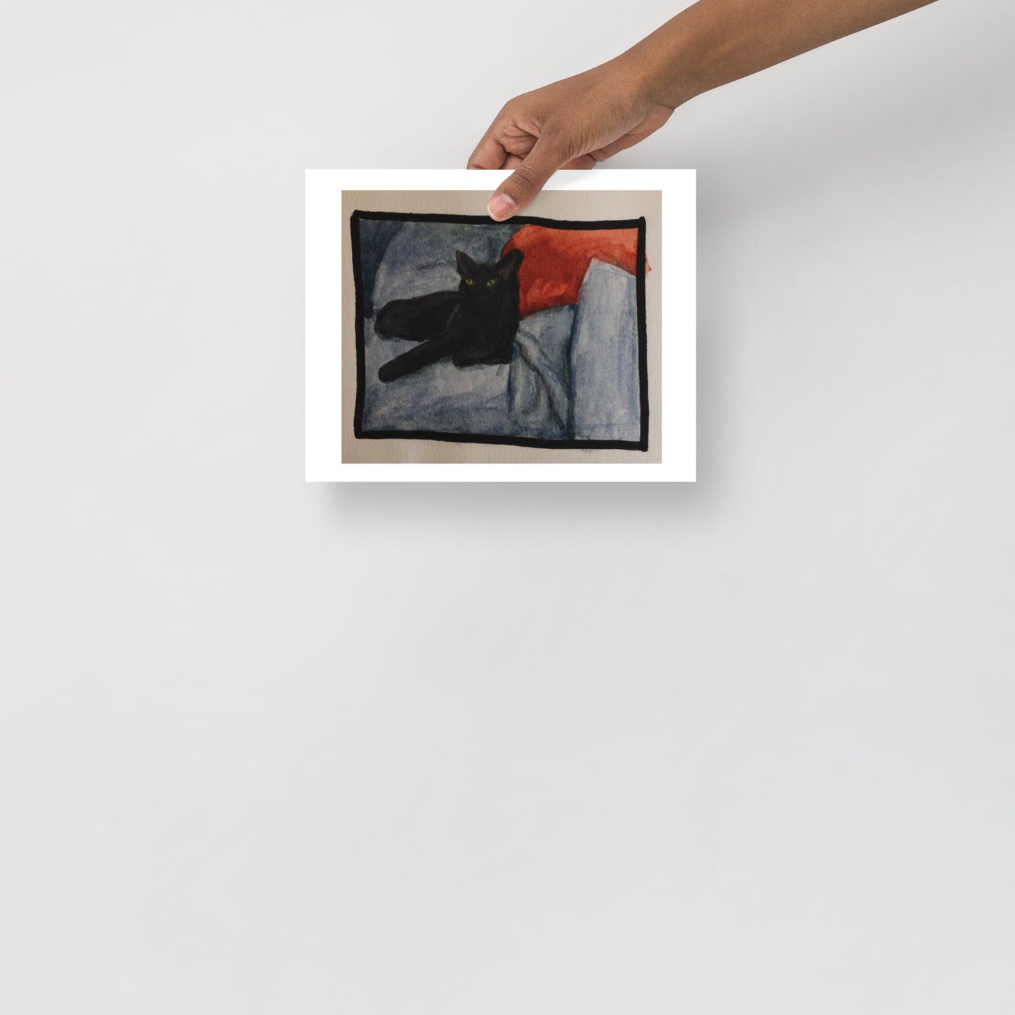 Product mockup of a watercolor painting that depicts a black cat lying on a textured blanket with a red pillow